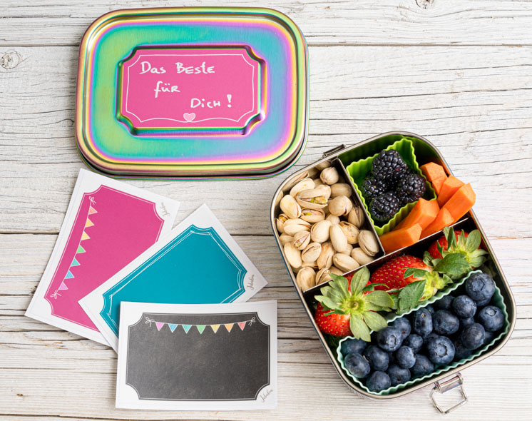 Bunte Edelstahl-Bentobox Rainbow mit Beeren, Nüssen und Snacks, unterteilt mit Bento-Cups, daneben Sticker und Lunchbox-Deckel mit Beschriftung.