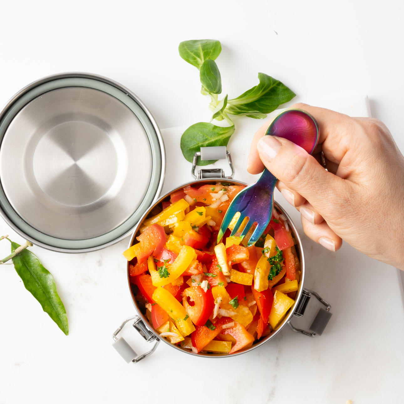 Mensch nutzt einen LEKKABOX Mini Göffel aus Edelstahl, um einen frischen Paprika-Salat aus einer kleinen Edelstahl Lunchbox zu essen – ideal für unterwegs.